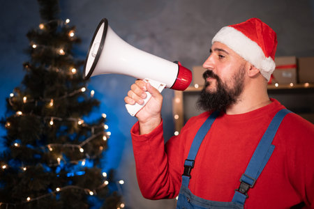 Small and medium business startup concept A young start-up businessman, wearing a santa claus hat and overalls, takes stands in a home office, holds a loudspeaker and yells.の写真素材