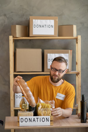 male volunteer packing donation box with food items of staple products on wooden table. Person man receiving donation food box. Donate food delivery. Donations grocery canned food.の写真素材
