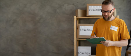 A male volunteer works at a donation center for the poor. Counts and writes boxes of donations. Humanitarian aid concept. Bannerの写真素材