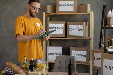 young volunteer man in t-shirt with volunteer inscriptions working in charity center on donation project indoors. calculating, sorting and packing items in donate boxesの写真素材