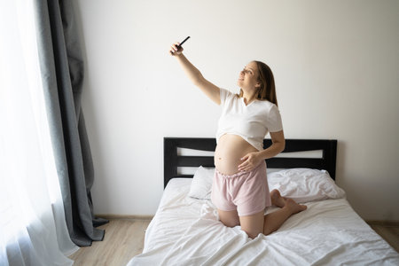 Pregnant woman with smart phone at home on bed, happy pregnancy selfie. Enjoying pregnancyの写真素材