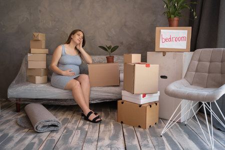moving to a new apartment. young pregnant woman with cardboard boxes sitting on sofa, housing programの写真素材