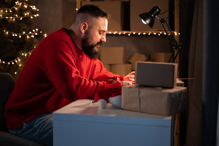 good earnings during the christmas sale. A young bearded Indian male entrepreneur sits at a table at night in a red suit checks orders on the site using a smartphone.の写真素材