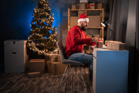 Young beginner bearded hindu man entrepreneur, small business owner in santa claus costume tired working at night in home office, tying red bow on delivery box. christmas sale.の写真素材