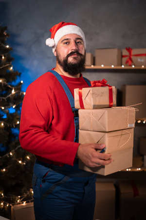 Small and Medium Business Startup Concept A young start-up businessman, wearing a santa claus hat and work overalls, takes a stand in a home office holding a lot of boxes in his hands.の写真素材