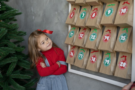 Handmade advent calendar hanging on the wall, girl standing near craft bags with sweets, holiday joy for kids, winter traditionsの写真素材
