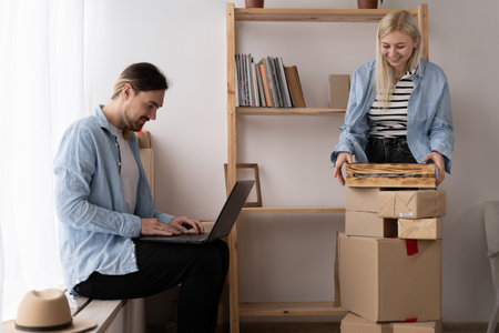 Young couple moving in new home. Happy homeowners sitting in a apartment using a laptop. A woman is unpacking things. Searching home decorating ideas on a notebookの写真素材