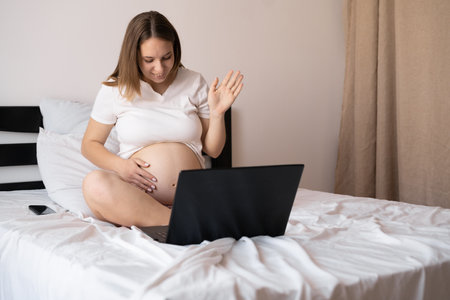 smiling pregnant woman sitting on bed with laptop computer. pregnancy, motherhood, internet and technologyの写真素材