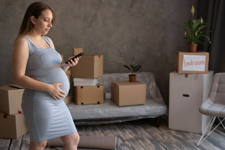 pregnant woman standing at new home during relocation using a phone for call in delivery serviceの写真素材