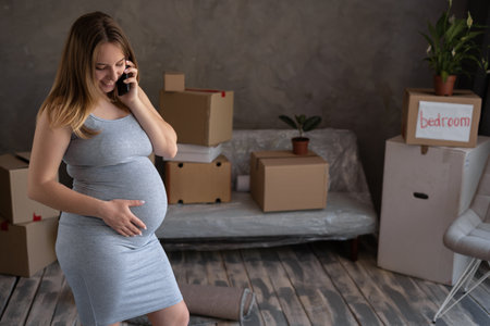 pregnant woman with belongings at new home using smartphone making call to delivery service, housing program for motherの写真素材