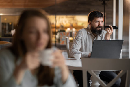 Bearded businessman working in cafe uses laptop and mobile phone voice assistant, blurred girl foreground, selectiveの写真素材