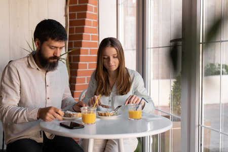 cheerful young couple out for breakfast in a cafe before work, sitting near window and enjoying conversation. woman and her boyfriend talking and laughing during mealの写真素材