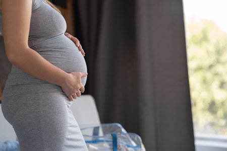 Close up of young pregnant woman with big tummy. Expecting mom standing indoors, holding belly, feeling unborn baby kicks. Pregnancy, motherhood, childbirth planningの写真素材