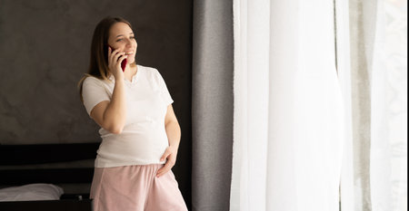 Pregnant woman talking on mobile phone while standing in living room at home with warm light, bannerの写真素材