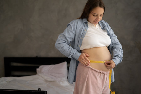Pregnant woman measuring her belly while standing against a grey background at homeの写真素材