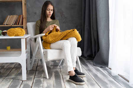 Young woman knitting a warm sweater from woolen threads while sitting in an armchair, hobby and needleworkの写真素材