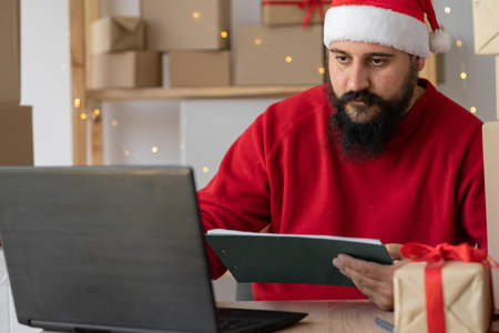 Happy Indian Santa Claus in hat using laptop while sitting at home table in workshop on Christmas Eve. online shopping sales on christmas holiday e-commerce websiteの写真素材