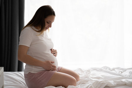 Attractive pregnant woman sitting in bed and holding her belly, bedroom window background. Last months of pregnancy.の写真素材