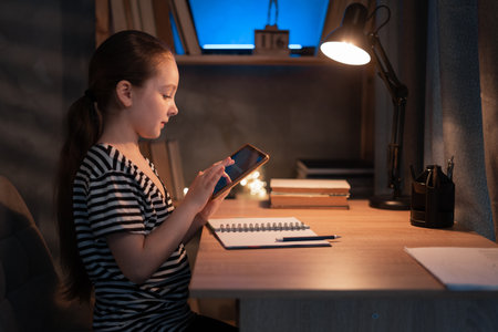Portrait of schoolgirl using doing homework writing in notebook sitting at desk at evening, holding and using tablet pad computer, enjoying distance homeschooling. Children and educationの写真素材