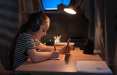 Happy teen girl at digital tablet doing homework online sitting at table at home in dark. Distant education, remote learningの写真素材