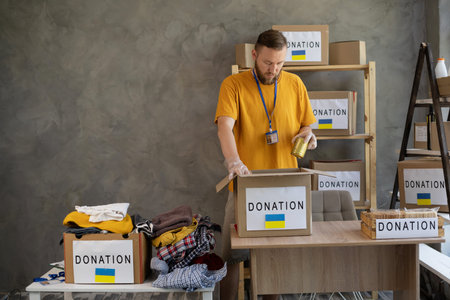 Young man wearing volunteer uniform working at charity center, packing donation boxes. Social help,...
