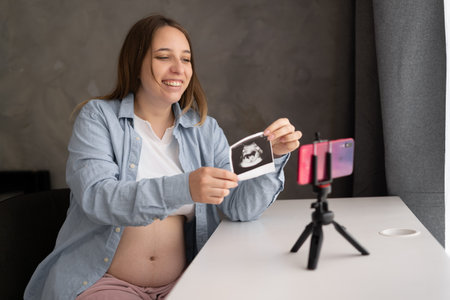 pregnant woman blogger showing ultrasound picture at camera, sitting at home, young future mom...
