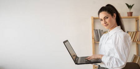 Successful hispanic millennial woman using and typing on laptop in modern office looking at camera....