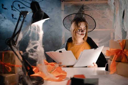 Dropshipping owner in Halloween makeup and witch costume looking at holiday sale paper report. Entrepreneur woman working at home office. Online marketing. Banner.の写真素材