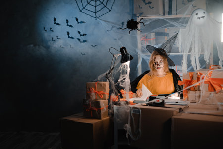 Dropshipping owner in Halloween makeup and witch costume reviewing paper sales report. Entrepreneur woman working at home office. Online marketing, discounts and promotions. Banner.の写真素材