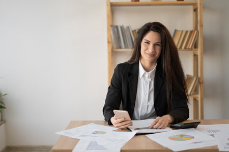 Latin businesswoman watching on smartphone during work at office. Idea of business and entrepreneur lifestyle. Young woman sit at desk with paper documents and cell phone. modern successful woman.の写真素材