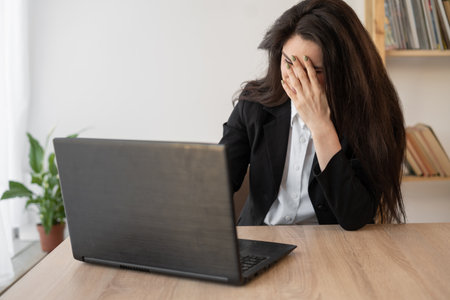 Woman working on laptop in office having a headache, migraine, relieving stress, chronic pain, help to calm. chronic fatigue syndromeの写真素材