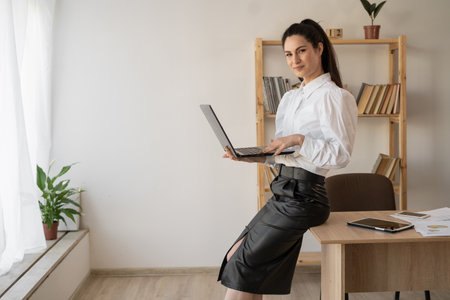 Shot of an attractive businesswoman working on a laptop at her workplace while sitting on a desk at office.の写真素材