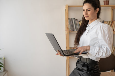 Successful hispanic millennial woman using and typing on laptop in modern office looking at camera. Female in businessの写真素材