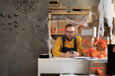 Small business owner man using mobile app on smartphone while checking parcel. Warehouse worker, seller in eyeglasses holding a phone. Big sale discounts and promotions in honor of Halloween.の写真素材