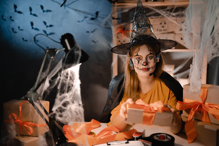 Startup small business entrepreneur or freelance caucasian woman in halloween makeup sitting at table among boxes holding out one with orange bow. online marketing packaging box and shippingの写真素材