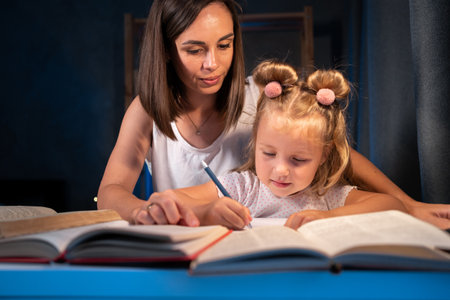 Happy little girl doing homework with mother at home at night. Young mother teaching small daughter to drawing, reading and writing develop her daughter skill. education, family and learningの写真素材