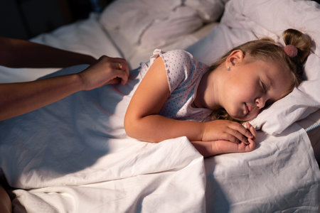Caring parent mom put blanket on sleeping little girl daughter on bed in dark bedroom at night. Loving mother take care asleep small kid. Parenting activity at home.の写真素材