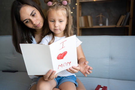 Happy mothers day. Child daughter congratulates mom and gives her postcard. Mum and girl smiling and hugging on sofa. Family holiday and togethernessの写真素材