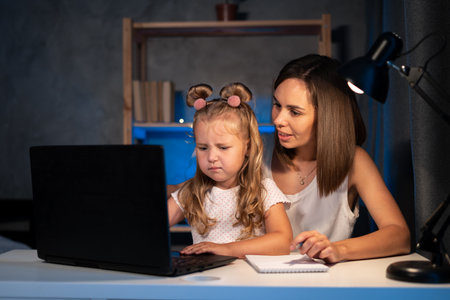 Woman with girl doing homework on laptop in the evening. little girl learning internet online class and do homework by using computer with mother help. family, e-learning and technologyの写真素材