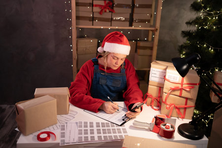 Woman in santa claus hat owner holds a package in a brown box, ready for delivery to a customer who orders online. Working at the home office on Christmas. SME, e-commerceの写真素材