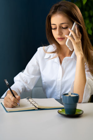 Young business woman sitting at table in cafe, talking on cell phone while taking notes in notebook. woman freelancer noting information for planning project job.の写真素材