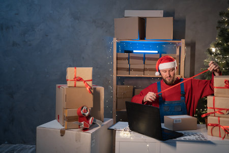 salesman in a Santa Claus hat takes an order by laptop while sitting in an office with boxes of gifts and surprises for Christmas for office workers. Shopping and delivery. Small businessの写真素材