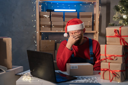 Start up. Depressed, stressed. Young Santa Claus working with laptop and delivery packaging box on table, small business owner working at home office, SME, Christmas shopping onlineの写真素材
