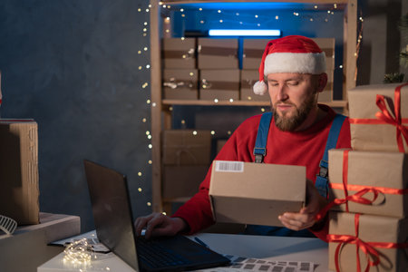 Santa Claus wearing hat holding gift box using laptop computer sitting at home office table late on Christmas eve. Ecommerce website xmas holiday online shopping e commerce saleの写真素材