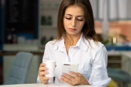 Charming young woman reading good news on mobile phone while relaxing in cafe, happy girl looking at photos on cell phone in free time in coffee houseの写真素材