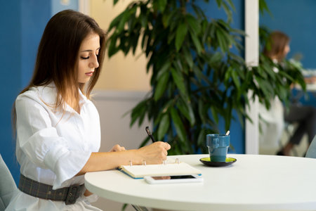 Happy young woman sitting at cafe working remotely and making note in planning notebookの写真素材