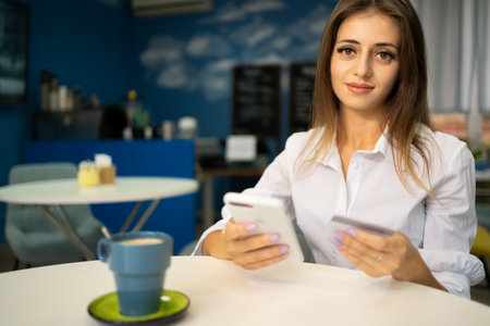 Beautiful young girl with a cheerful expression holding a smartphone and a credit card, banking online or shopping while in a cafe interior. payment and vacationの写真素材