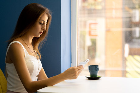 Beautiful cute young businesswoman in cafe using mobile phone and drinking coffee smiling near windowの写真素材