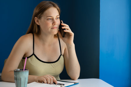 Young woman calling with cell telephone while sitting alone in coffee shop during free time, having talking conversation with mobile phone while rest in cafeの写真素材