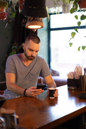 Successful young businessman is using a smartphone and drinking coffee while sitting at bar counter in a modern urban cafeの写真素材